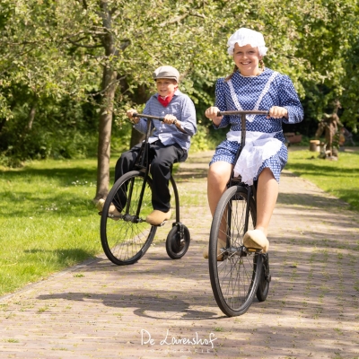 De afbeelding toont twee kids op een ouderwetse fiets in klederdracht aan het spelen
