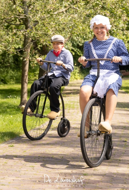 De afbeelding toont twee kids op een ouderwetse fiets in klederdracht aan het spelen